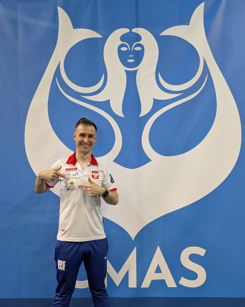Bartosz Jakubowiak pointing to the Freediver.club logo on his Polish national-team jersey in front of the CMAS banner in Novi Sad